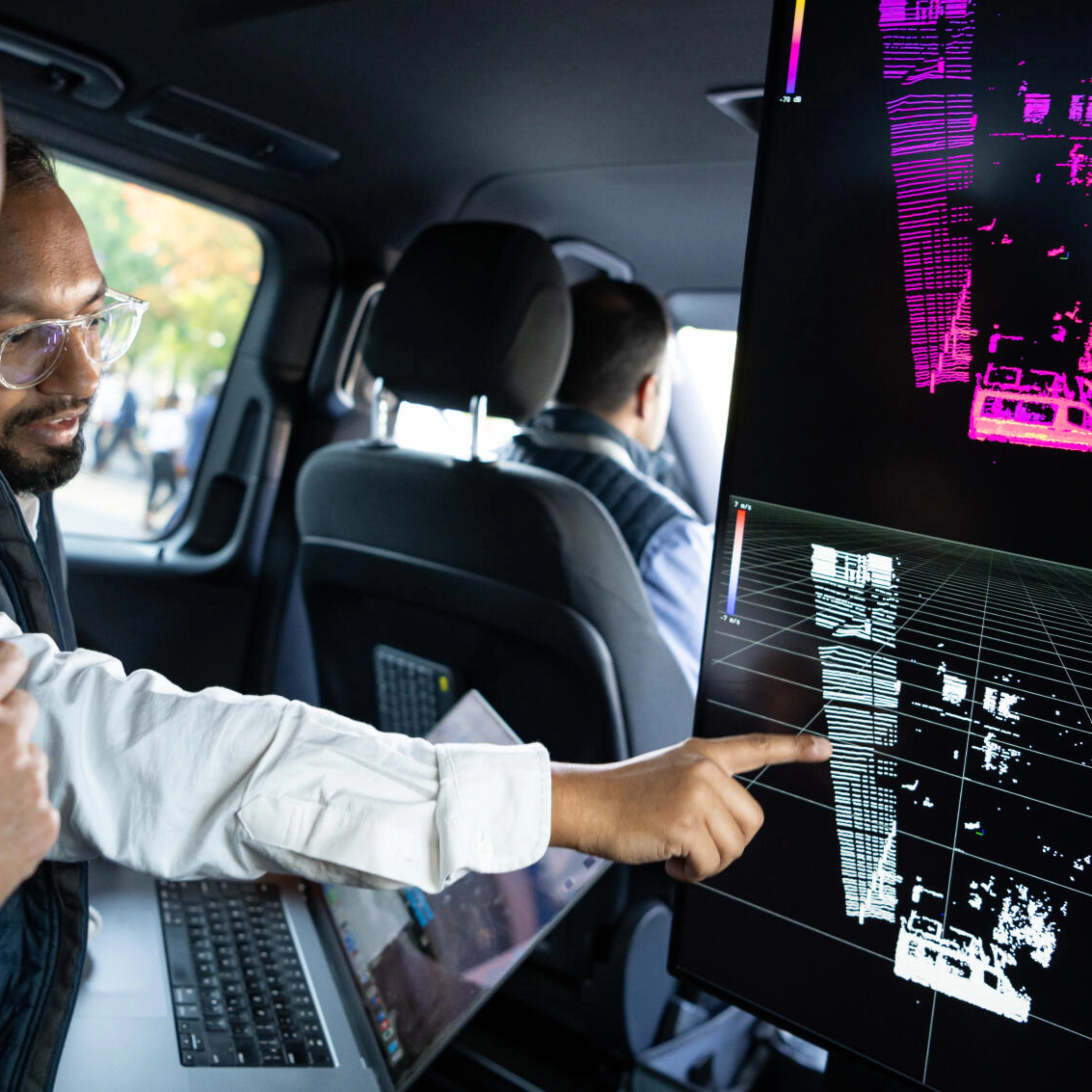 Two men in a car analyze 3D sensor data on a large screen for autonomous driving.