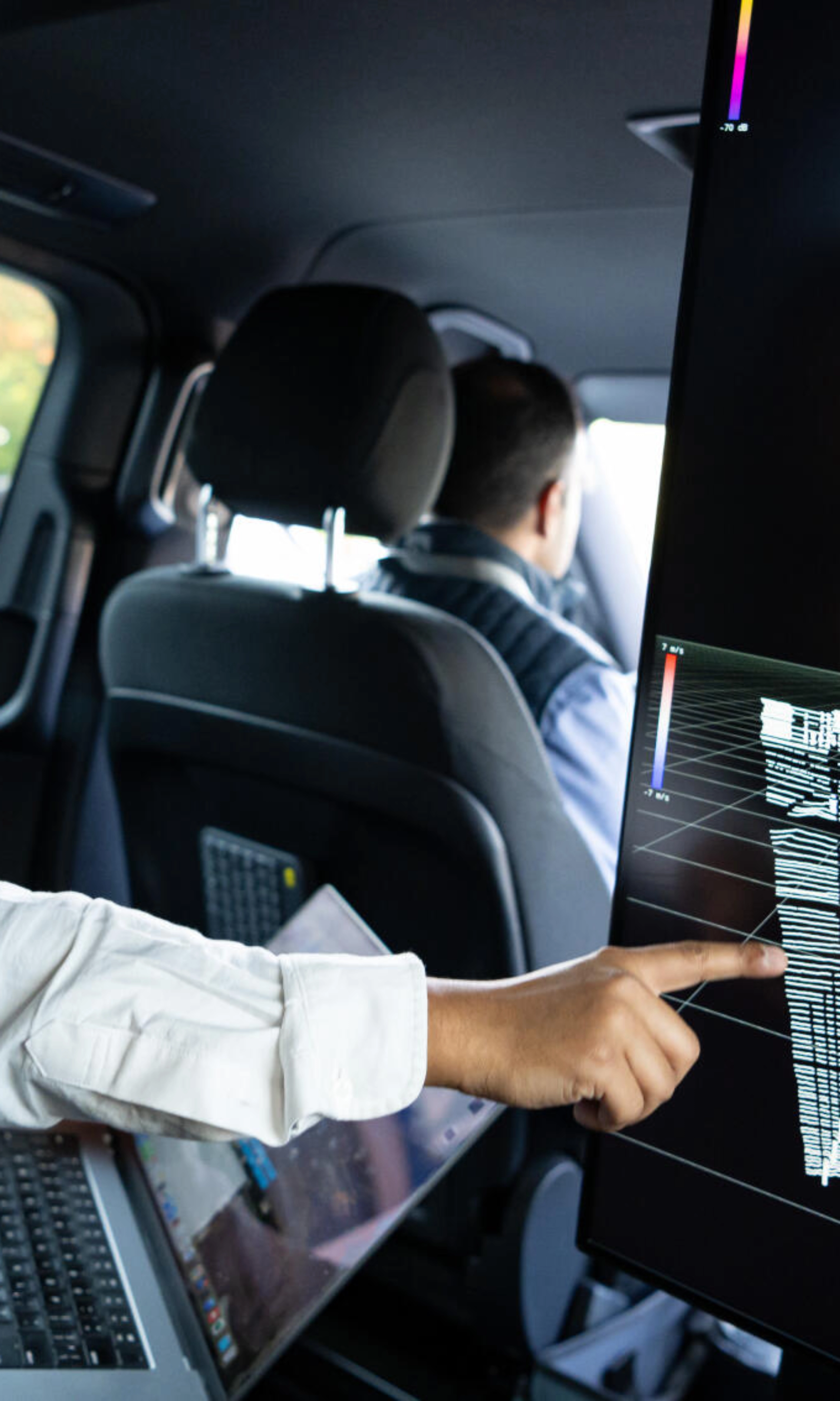 Two men in a car analyze 3D sensor data on a large screen for autonomous driving.