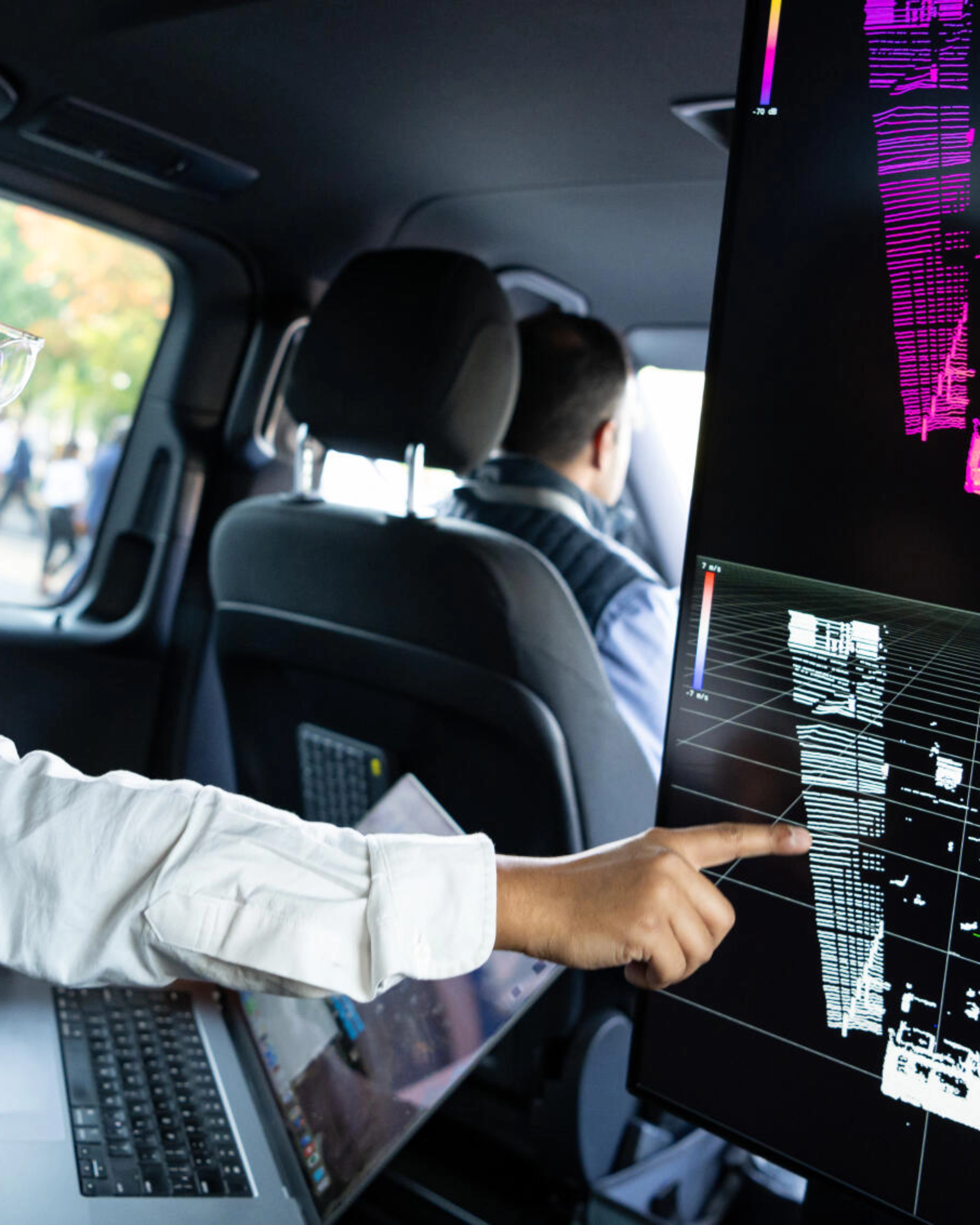 Two men in a car analyze 3D sensor data on a large screen for autonomous driving.