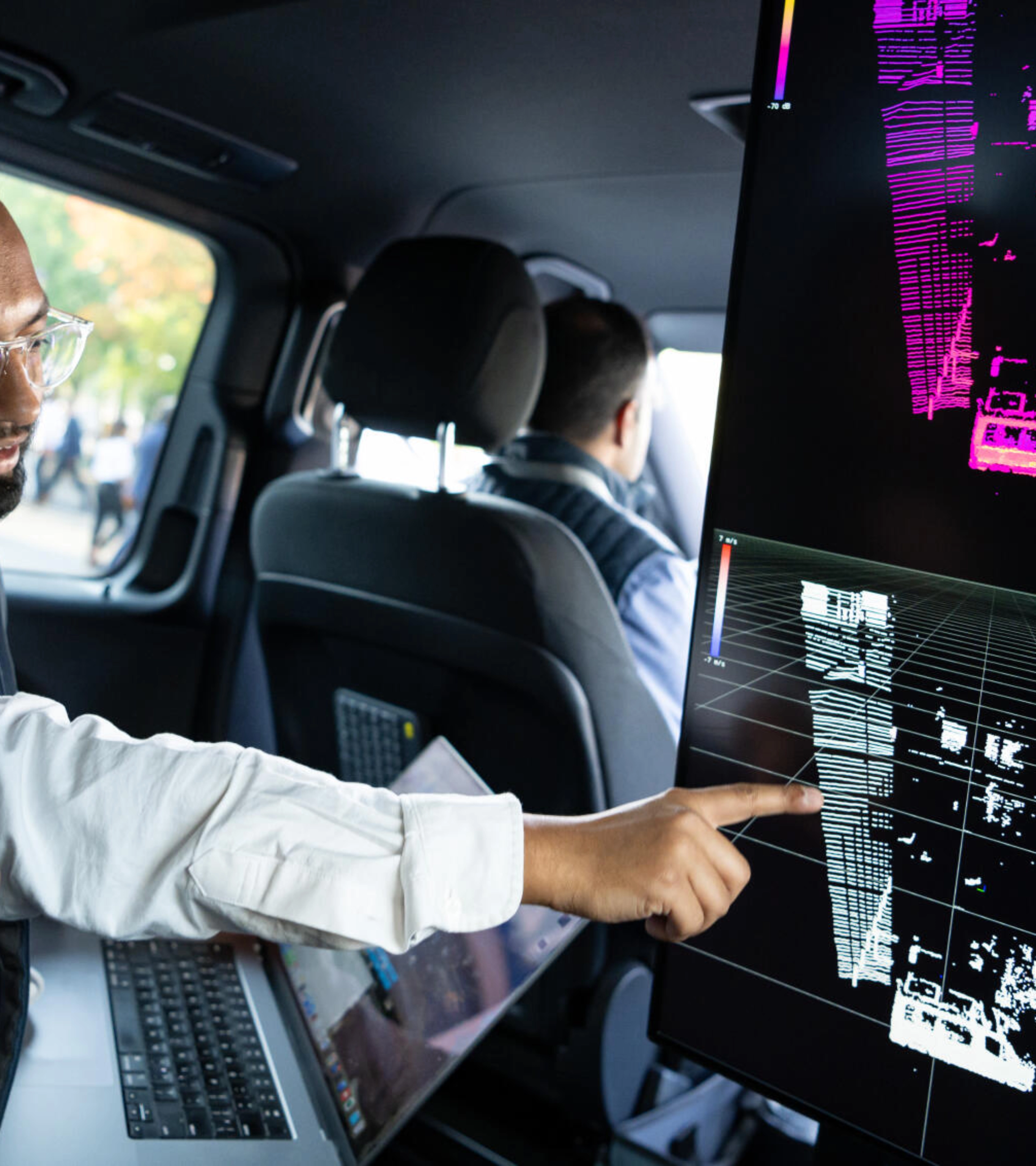 Two men in a car analyze 3D sensor data on a large screen for autonomous driving.