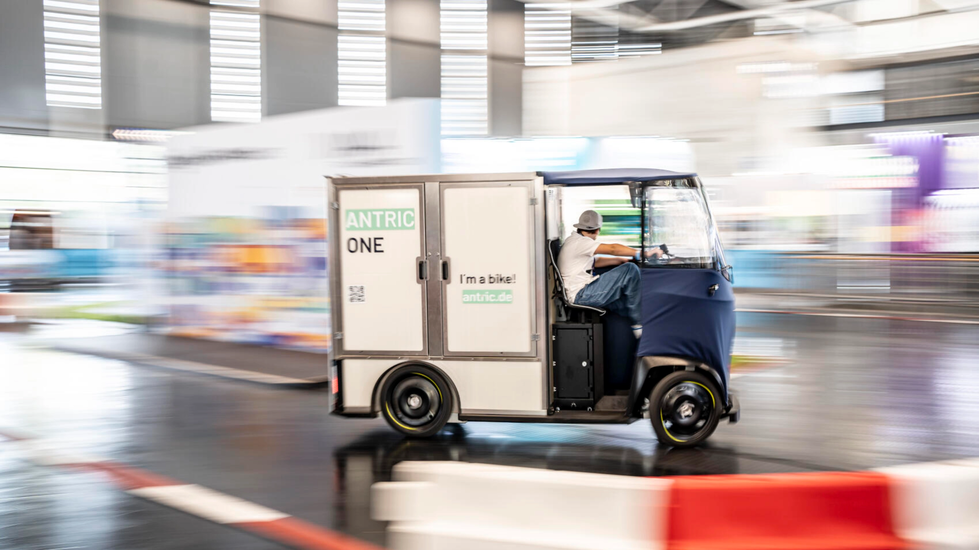 Small cargo bike vehicle labeled ANTRIC ONE driving indoors at a trade fair.